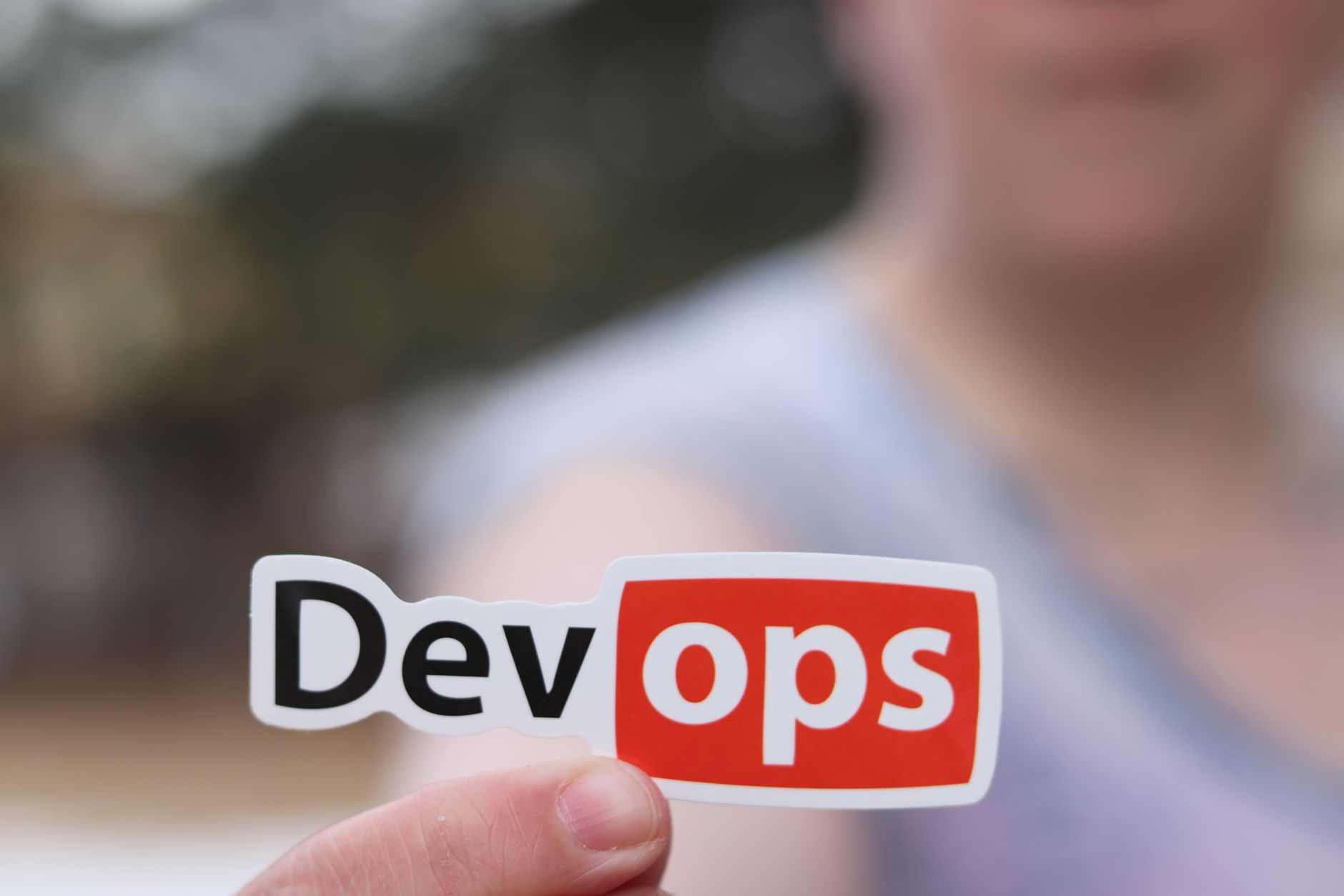 Dłoń trzymająca naklejkę z napisem DevOps na tle pleneru